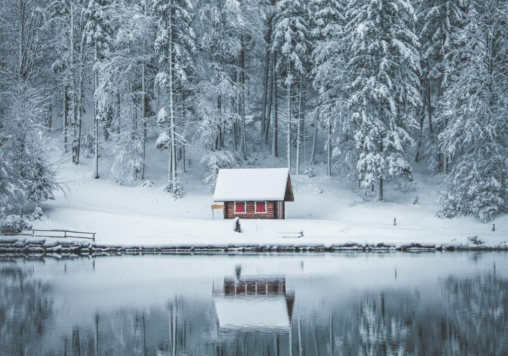 Tranquil cabin amidst snow-covered forest reflecting in the icy lake.